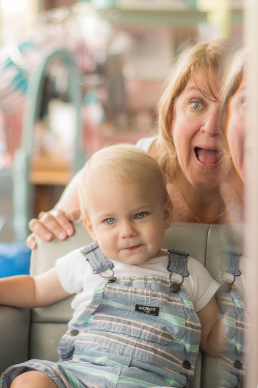 blue eyed toddler boy sitting in a chair looking in a mirror as his grandmother pops up behind him to make him smile